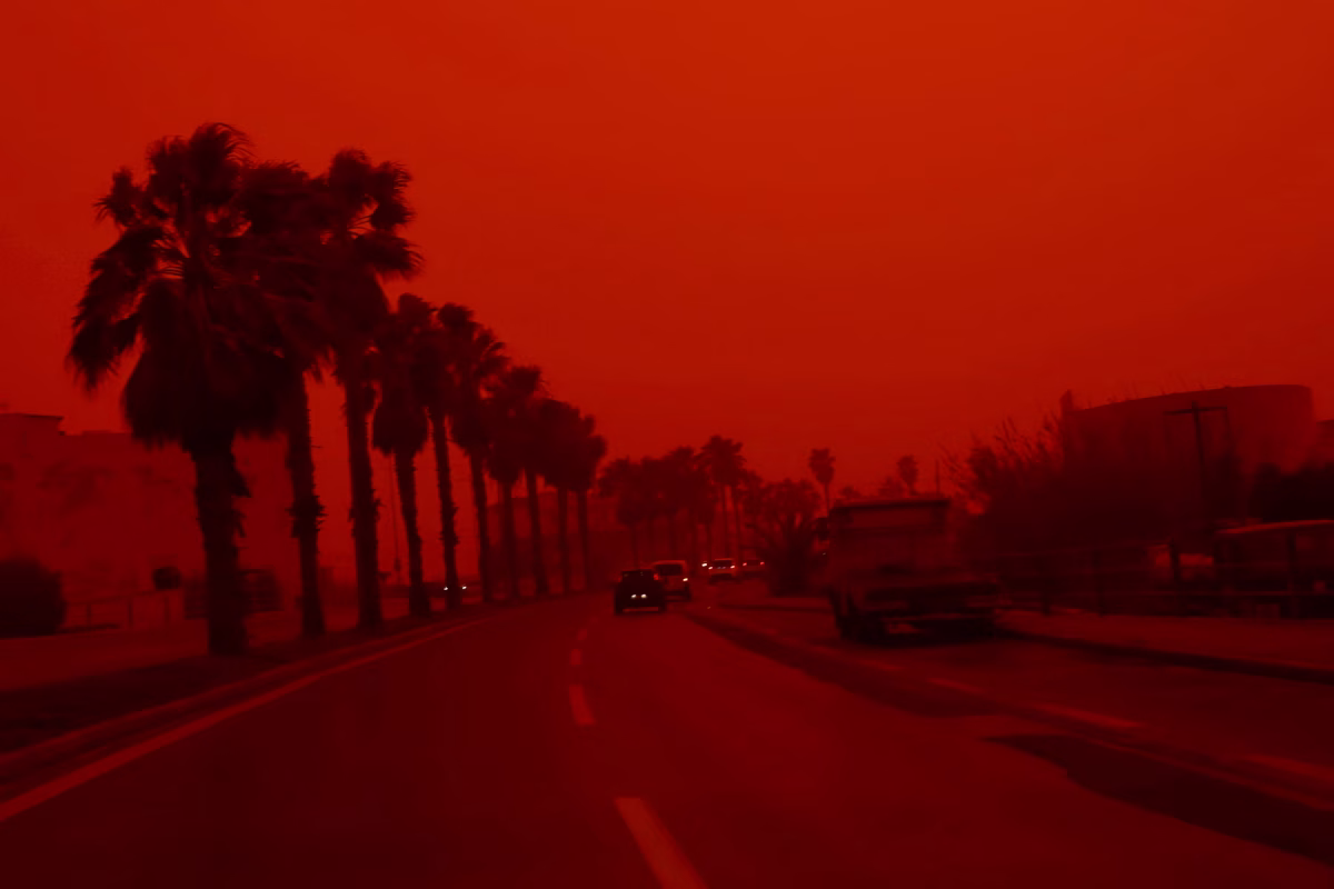 Vista panorâmica de uma cidade coberta por uma densa nuvem de poeira de coloração laranja intensa, com prédios parcialmente ocultos pela névoa.