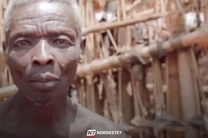 Homem idoso com aparência de eremita sentado em frente a uma cabana rústica e isolada na floresta.