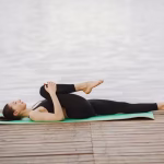 Pessoa deitada sobre um tapete de yoga praticando alongamentos leves em uma sala iluminada.