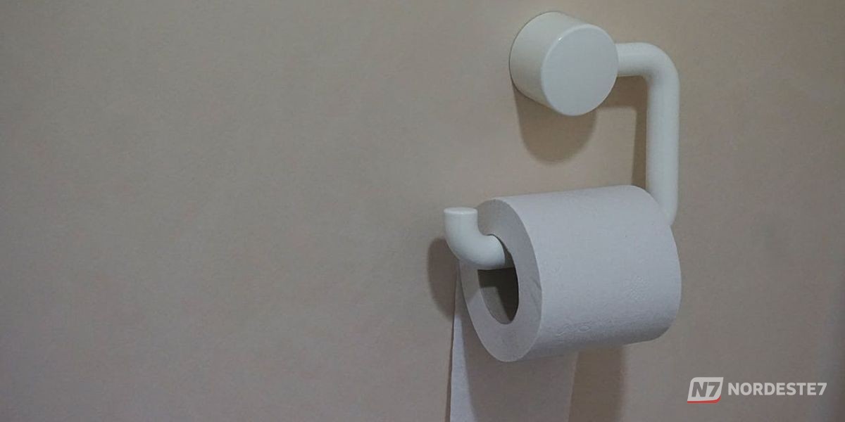 Mão segurando papel higiênico acima de um vaso sanitário com água, ilustrando o hábito de descarte que pode causar entupimento na rede de esgoto urbana.