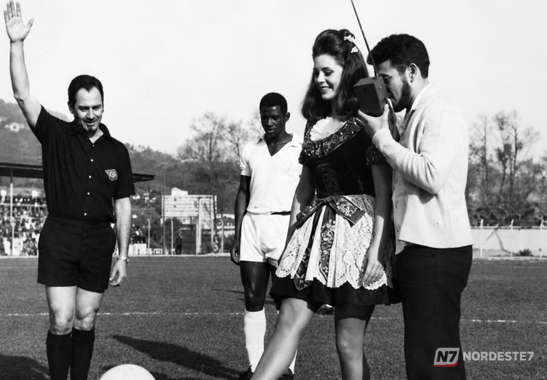 Fotografia histórica de uma atriz sorridente em um campo de futebol, prestes a dar o pontapé inicial de um torneio que leva o seu nome, cercada por jogadores e público.
