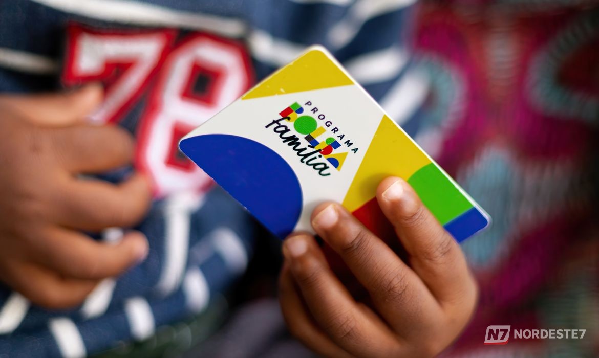 Cartão do Bolsa Família em destaque na mão de um beneficiário em frente a um posto de atendimento do Cadastro Único, simbolizando a atualização cadastral.