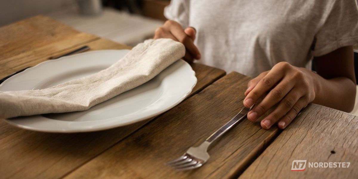 Close-up de um garfo de prata posicionado de forma precisa à esquerda de um prato de porcelana em uma mesa de jantar sofisticada.