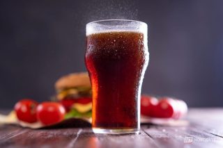 Copo de refrigerante com gelo ao lado de um prato de comida durante uma refeição, ilustrando o hábito de consumir a bebida com alimentos.