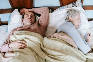 Pessoa deitada na cama com expressão de desconforto, esticando a mão para alcançar um copo de água na mesa de cabeceira, ilustrando o sintoma de boca seca ao acordar.
