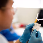 Frasco de vacina sendo preparado por profissional de saúde com luvas em um laboratório clínico na Inglaterra, representando a nova imunoterapia contra o câncer.