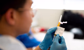 Frasco de vacina sendo preparado por profissional de saúde com luvas em um laboratório clínico na Inglaterra, representando a nova imunoterapia contra o câncer.