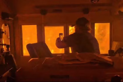 Trem de passageiros cruzando trilhos cercados por chamas intensas e fumaça de incêndio florestal causado por calor extremo.