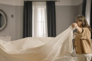 Mulher estendendo um lençol branco limpo sobre uma cama bem arrumada em um quarto iluminado.